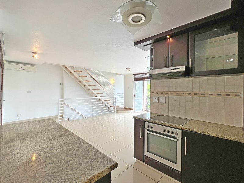 Open-plan living area featuring a modern kitchen with granite countertops, built-in oven, and hob, leading to a staircase and tiled living space.