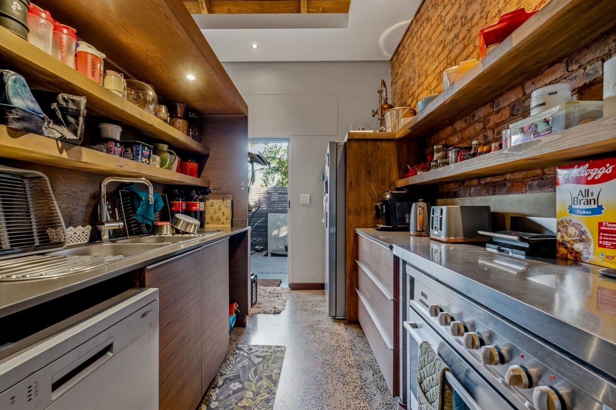 Modern industrial-style kitchen featuring exposed brick walls, stainless steel countertops, built-in oven, and polished concrete flooring with direct access to an outdoor area.