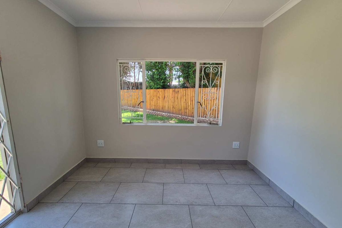 A bright, empty living room featuring neutral wall tones, large format floor tiles, and a window with decorative security bars overlooking the rear garden.