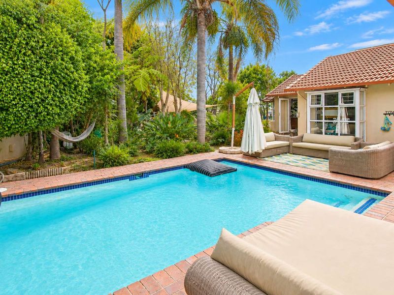A well-maintained outdoor pool area featuring brick paving, lush garden landscaping, and direct access to the home through classic french doors.