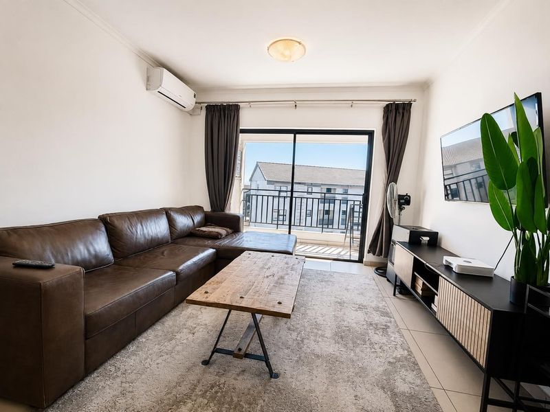 A bright living room featuring tiled flooring, a wall-mounted air conditioning unit, and a sliding glass door providing access to a balcony.