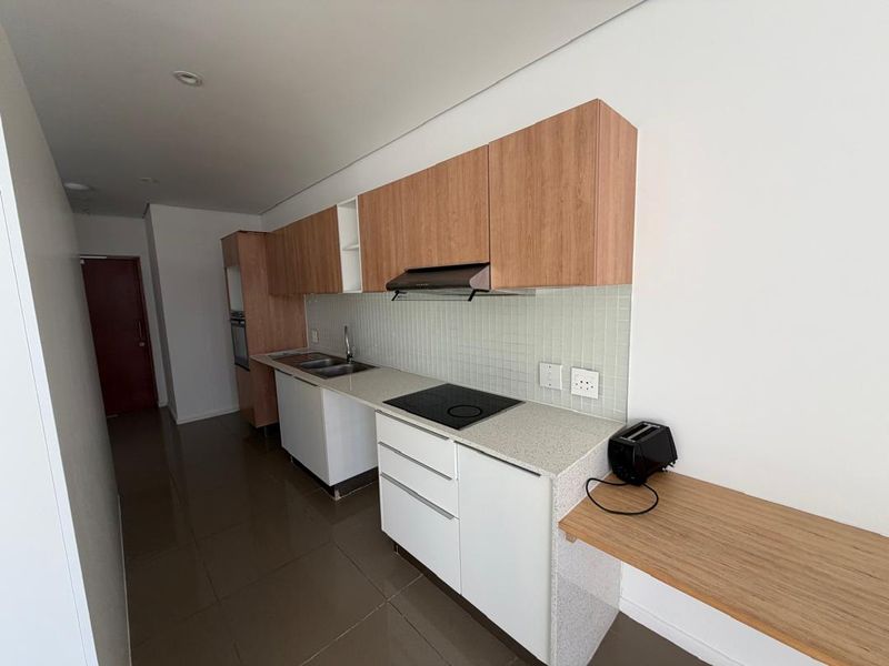 Modern galley-style kitchen featuring light wood-tone upper cabinetry, white lower cabinets, stone countertops, integrated cooktop, and a tiled backsplash.