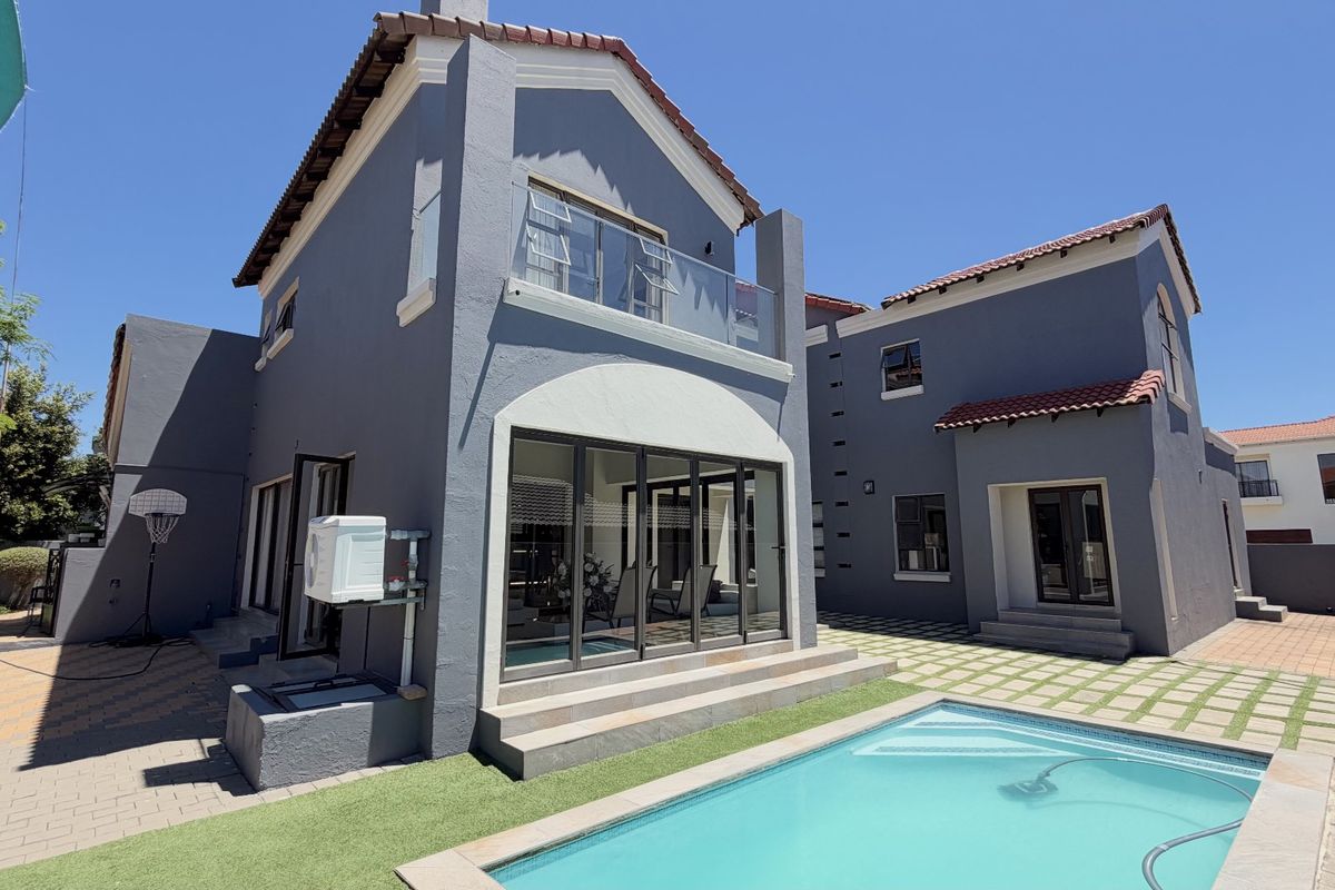Rear view of a modern two-story house featuring a swimming pool, paved patio area, artificial lawn, and a glass-railed balcony.