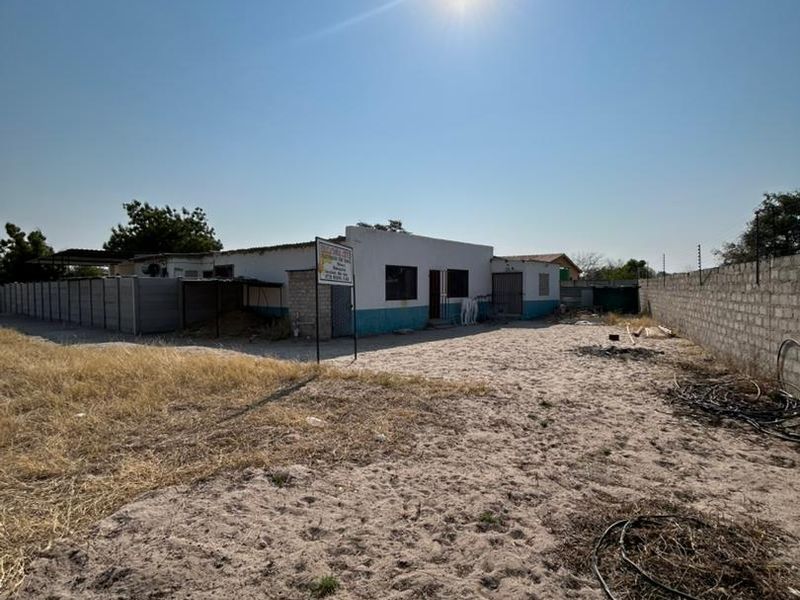 Front exterior view of a single-story house featuring a large sandy yard, concrete block boundary walls, and electric fencing.