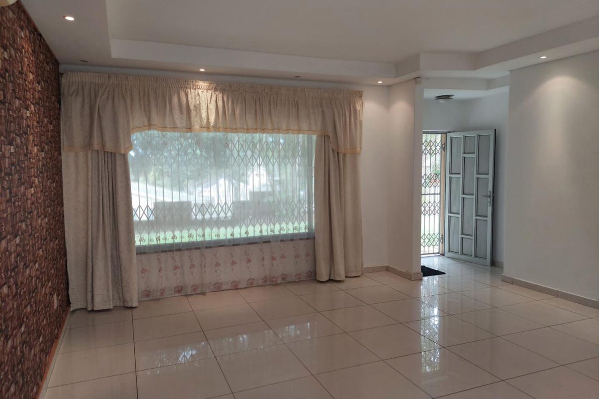 A spacious, empty living room featuring large windows with security gates, tiled flooring, recessed ceiling lighting, and a decorative textured feature wall.