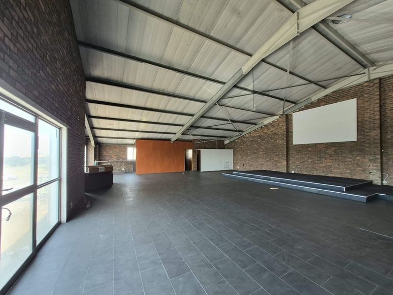 Spacious open-plan office area featuring exposed brick walls, a pitched metal roof, and a raised stage platform, offering a versatile industrial-style workspace.
