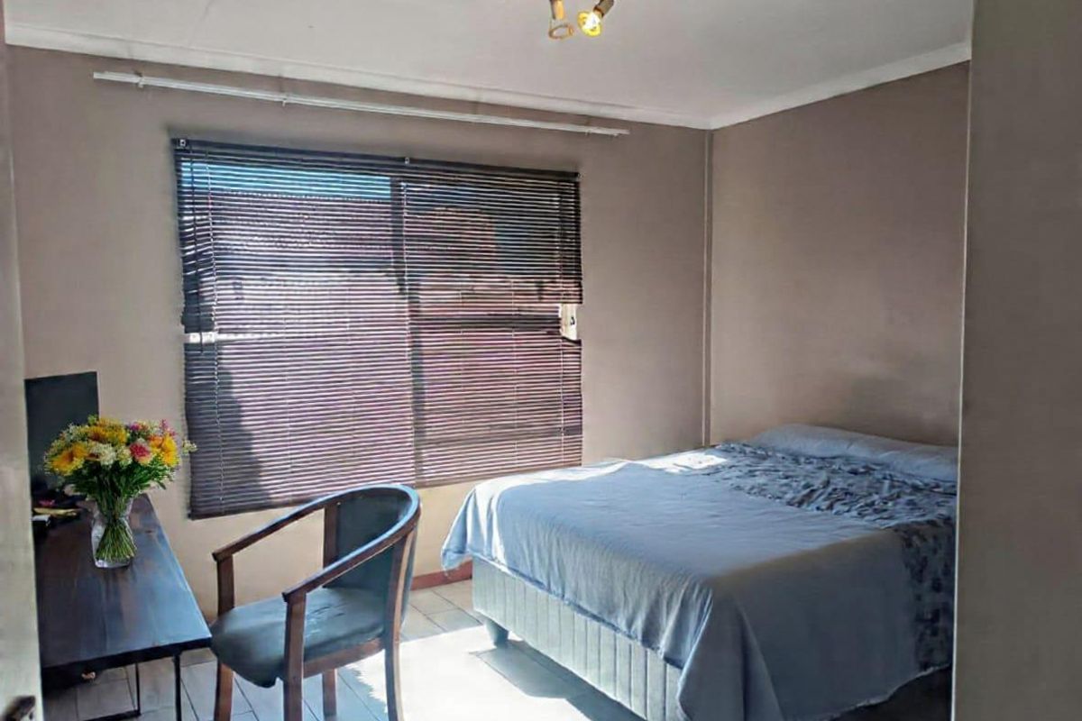 A functional bedroom featuring tiled flooring, a ceiling light fixture, and a window fitted with venetian blinds.