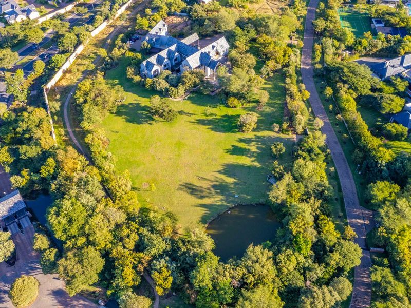An aerial view of a large residential plot featuring expansive green lawns, mature trees, and a private pond within a landscaped setting.