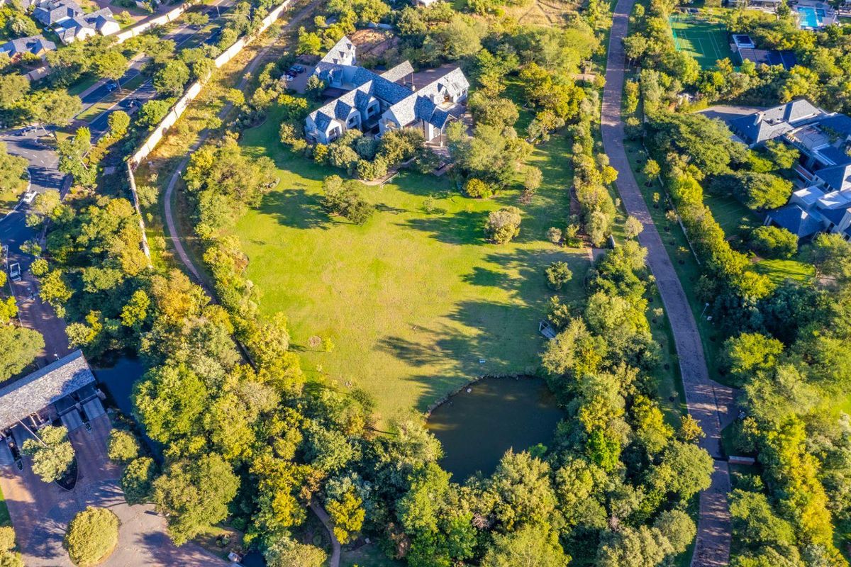 An aerial view of a large residential plot featuring expansive green lawns, mature trees, and a private pond within a landscaped setting.