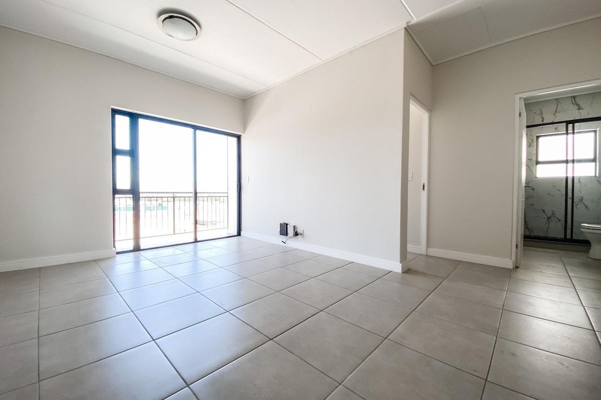 A bright, empty bedroom featuring neutral tiled flooring, a sliding glass door leading to a balcony, and an adjacent en-suite bathroom.