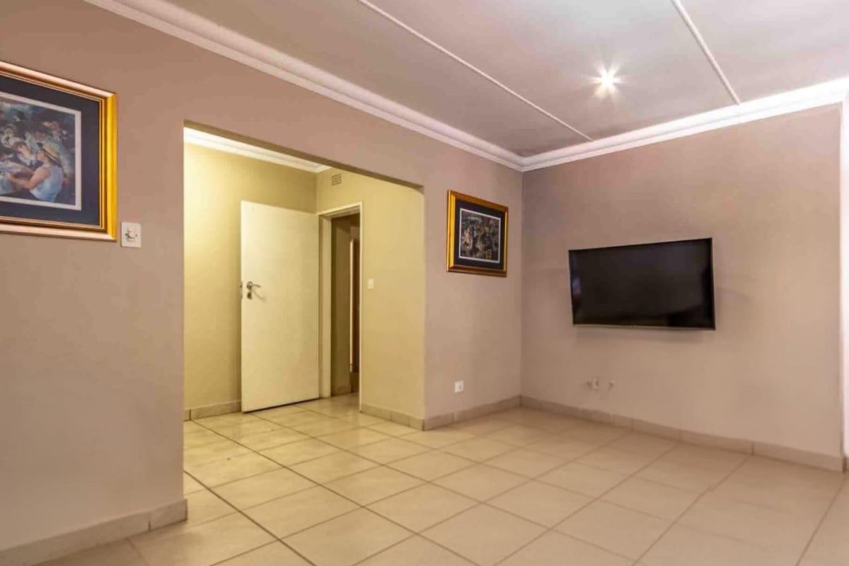 A spacious living room featuring neutral tiled flooring, coved ceilings, and a wall-mounted television point, leading into a connecting hallway.