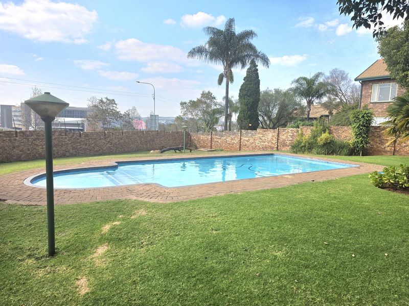 A well-maintained outdoor swimming pool area featuring a paved surround, established garden, and secure brick boundary wall.