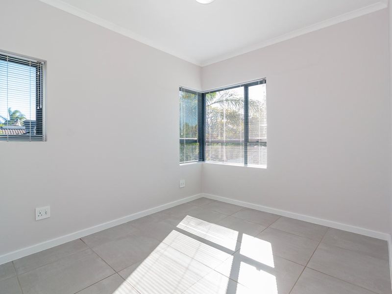 A bright, empty bedroom featuring neutral tiled flooring, corner windows with fitted venetian blinds, and clean, well-maintained wall finishes.