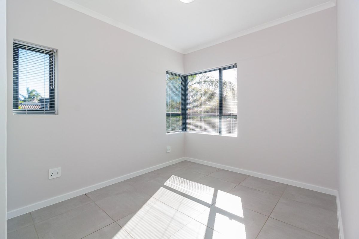 A bright, empty bedroom featuring neutral tiled flooring, corner windows with fitted venetian blinds, and clean, well-maintained wall finishes.