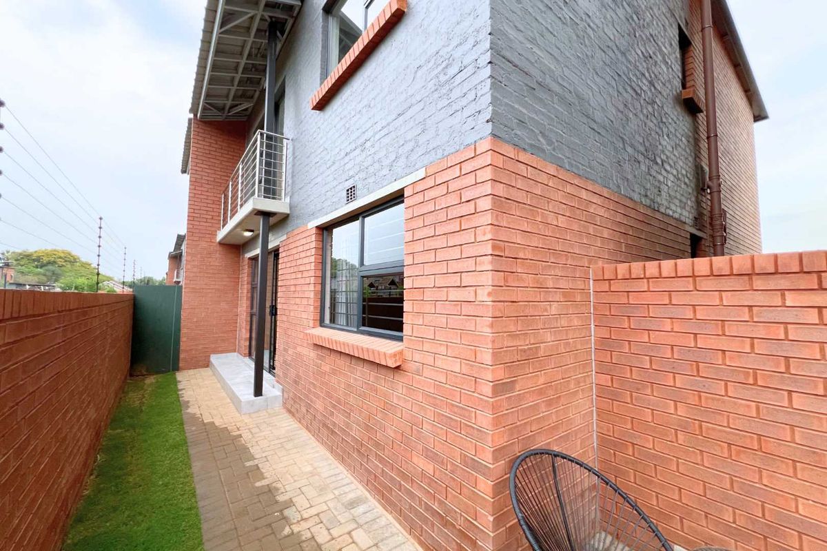 A view of the rear exterior of the apartment building, featuring face-brick walls, a small paved walkway, and a balcony on the upper level.