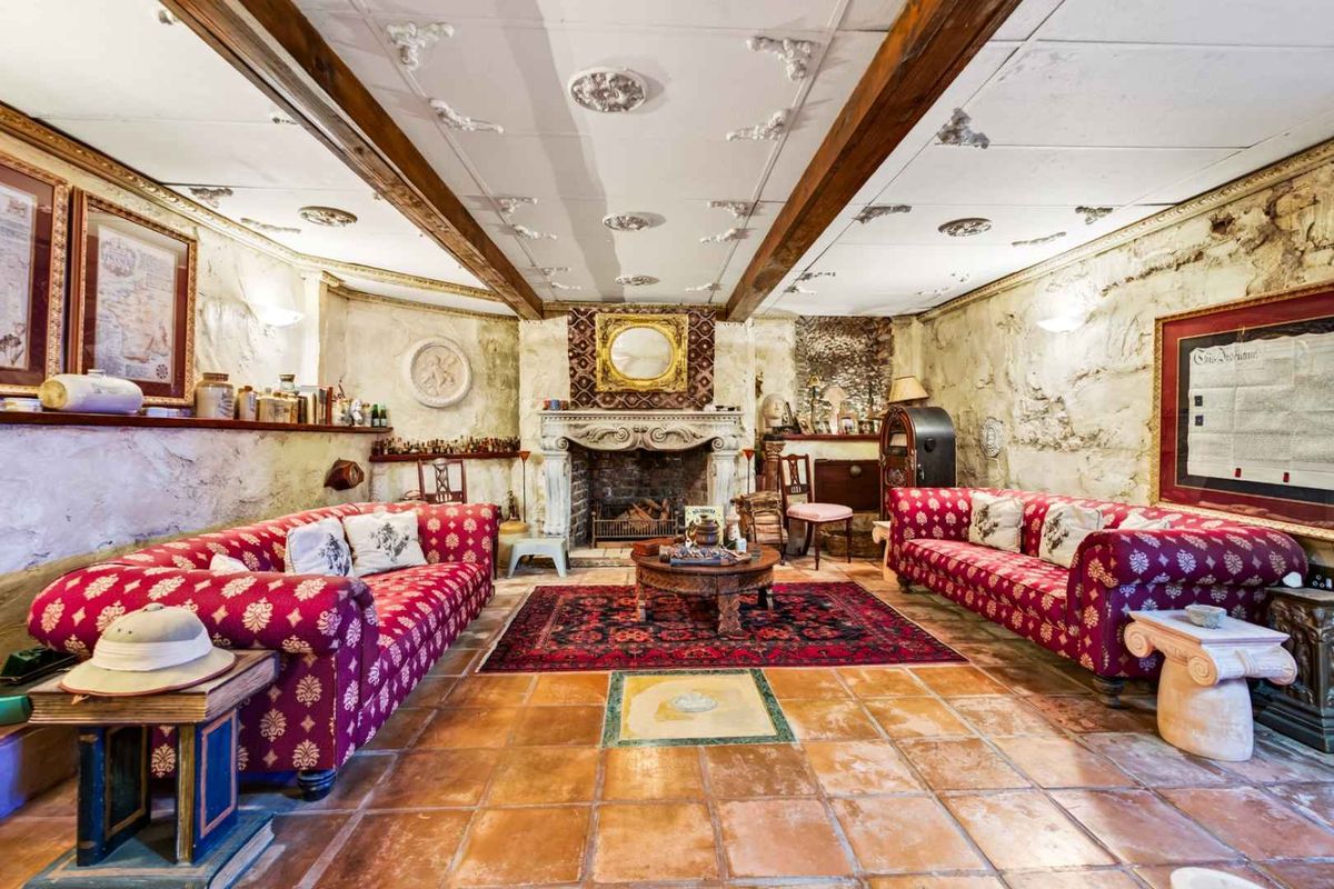 A rustic living room featuring exposed timber ceiling beams, textured stone walls, terracotta flooring, and a prominent stone fireplace.