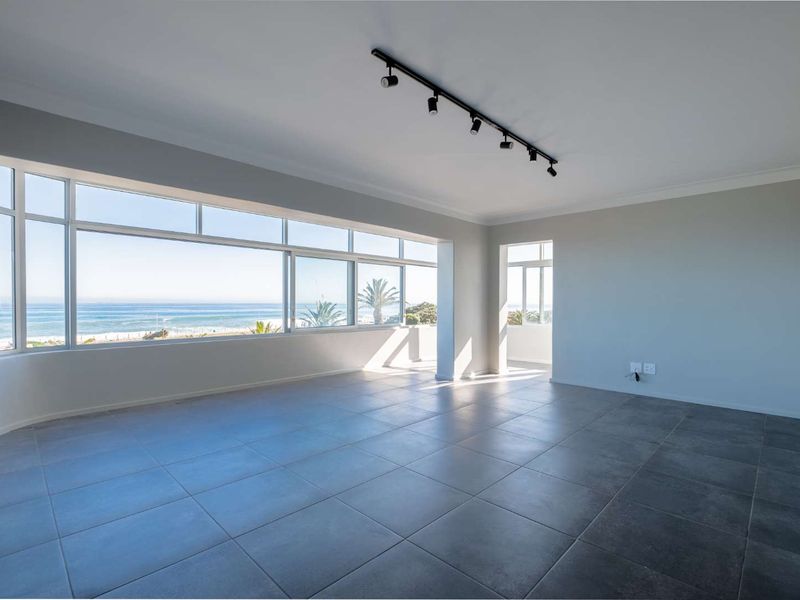 Spacious, light-filled living area featuring large format floor tiles, modern track lighting, and expansive windows offering panoramic ocean views.