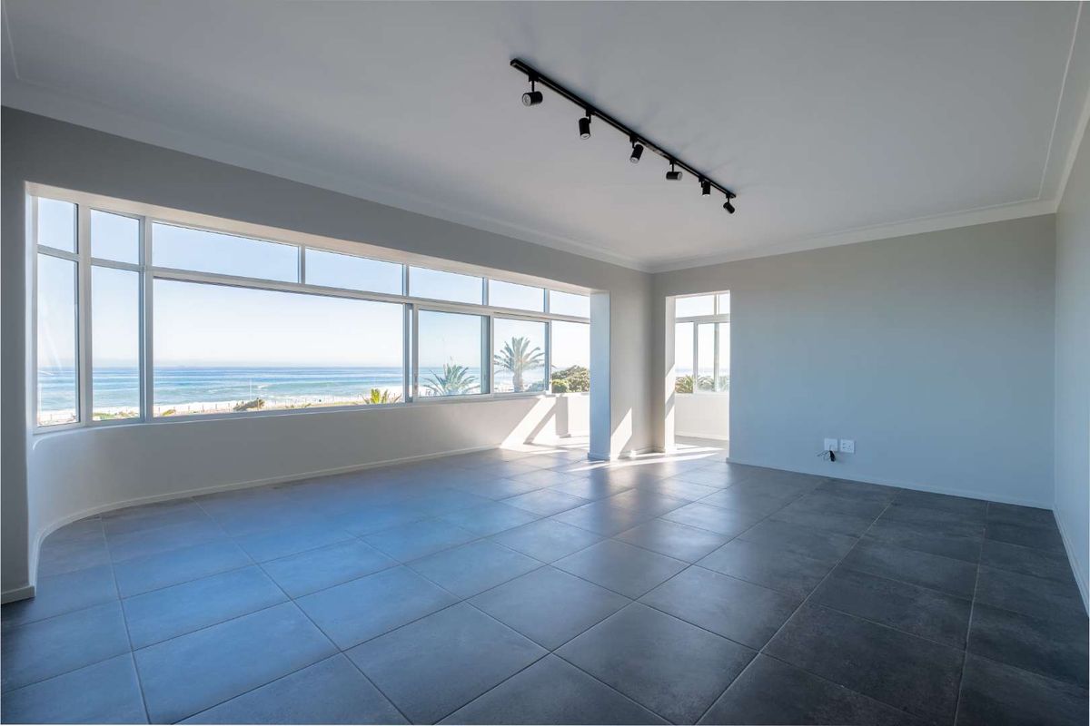 Spacious, light-filled living area featuring large format floor tiles, modern track lighting, and expansive windows offering panoramic ocean views.