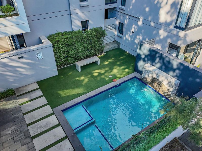 An elevated view of a private apartment courtyard featuring a swimming pool with a built-in water feature, artificial lawn, and paved walkways.