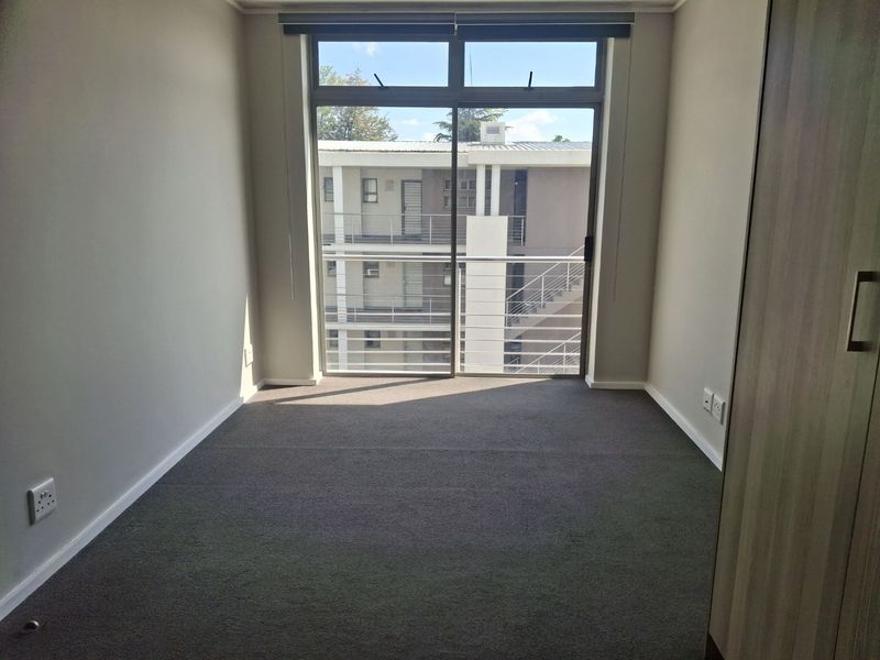 A bright, empty bedroom featuring neutral walls, carpeted flooring, a built-in cupboard, and a sliding glass door leading to a balcony.
