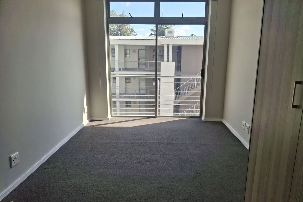 A bright, empty bedroom featuring neutral walls, carpeted flooring, a built-in cupboard, and a sliding glass door leading to a balcony.