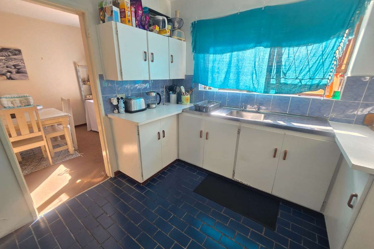 A functional kitchen featuring white cabinetry, blue tiled splashbacks, and dark floor tiles, with an adjacent dining area visible through the doorway.