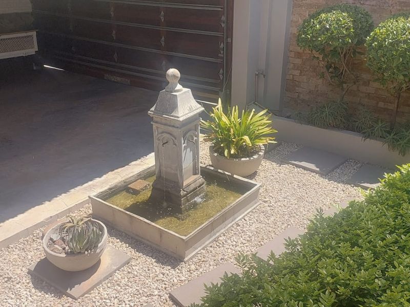 A well-maintained garden area featuring a central stone fountain, gravel landscaping, and manicured shrubs set against a brick boundary wall.