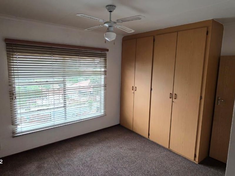 A bedroom featuring built-in wooden cupboards, a ceiling fan, venetian blinds, and a textured brick feature wall.