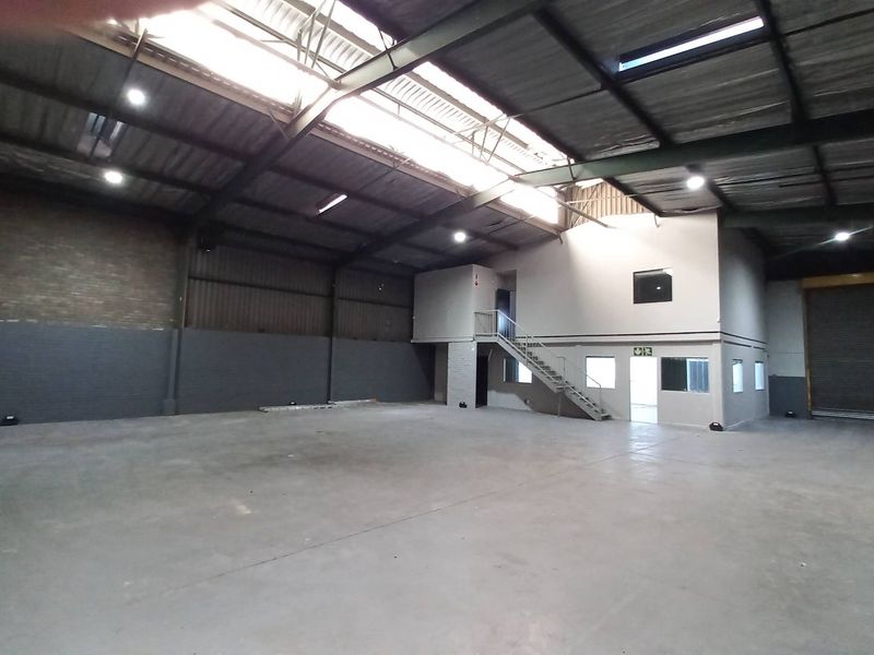 Spacious industrial warehouse featuring high ceilings, exposed steel trusses, concrete flooring, a mezzanine level with office space, and a large roller shutter door.