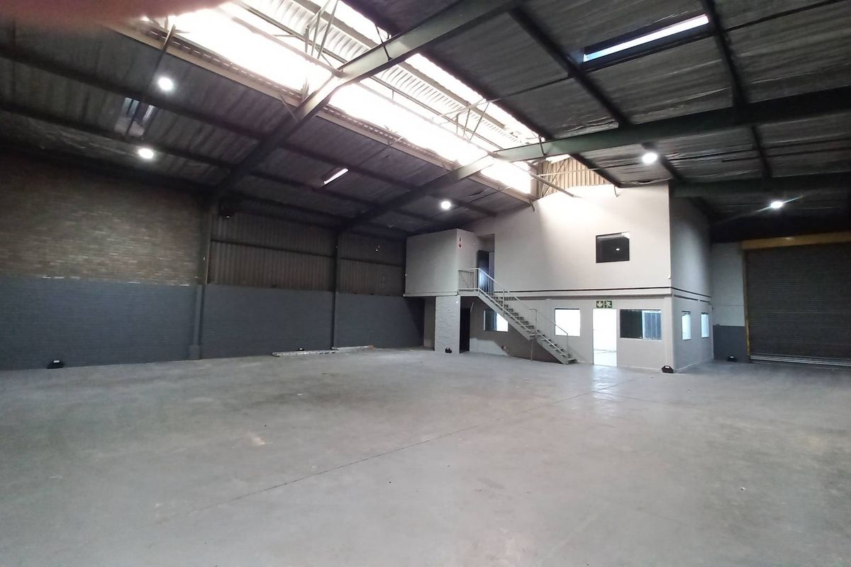 Spacious industrial warehouse featuring high ceilings, exposed steel trusses, concrete flooring, a mezzanine level with office space, and a large roller shutter door.