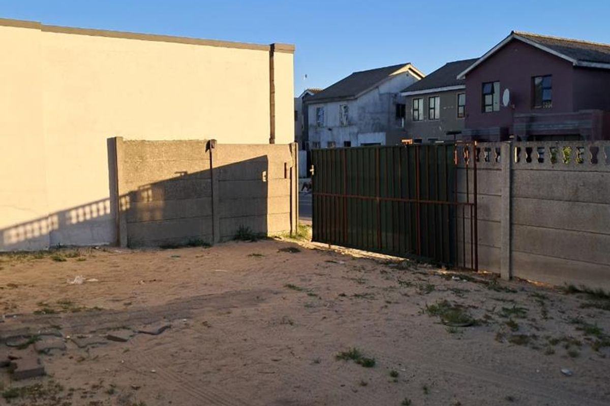 An unpaved rear yard area enclosed by precast concrete boundary walls and a metal gate, showing signs of neglect and lack of landscaping.