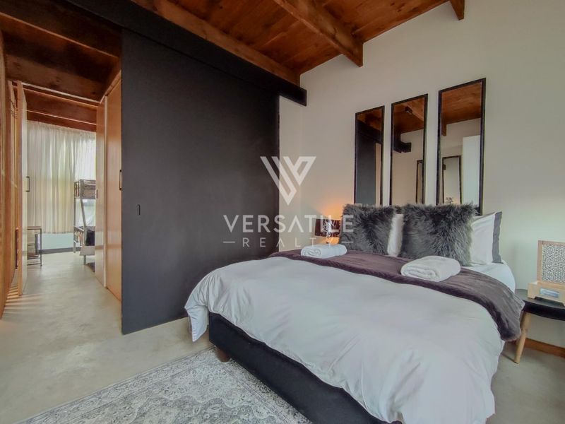 A modern bedroom featuring exposed timber ceiling beams, polished concrete floors, and a large sliding door leading to an adjacent space.