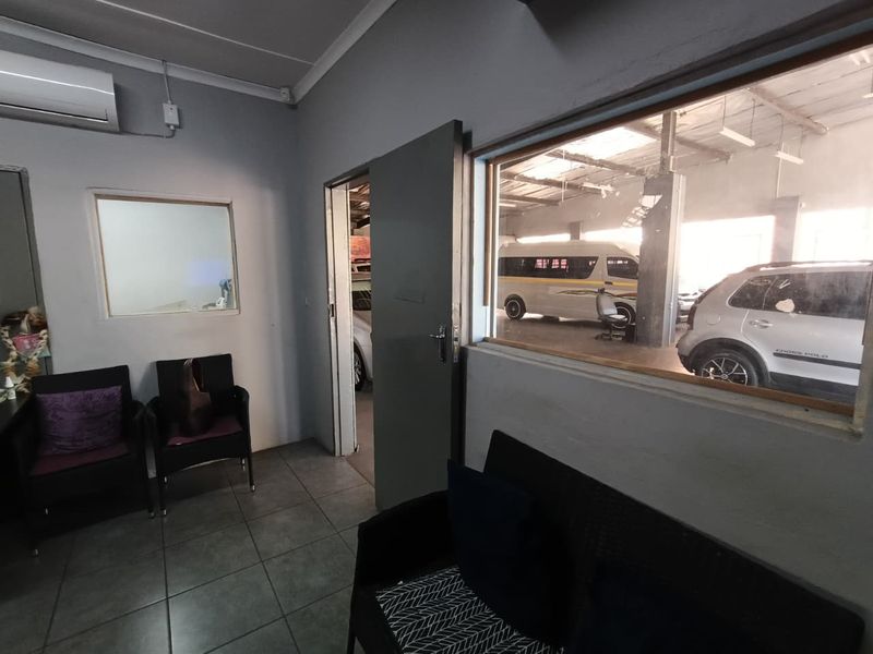 A functional office space featuring tiled flooring, a split-system air conditioner, and an internal window providing a view into the adjacent industrial workshop area.
