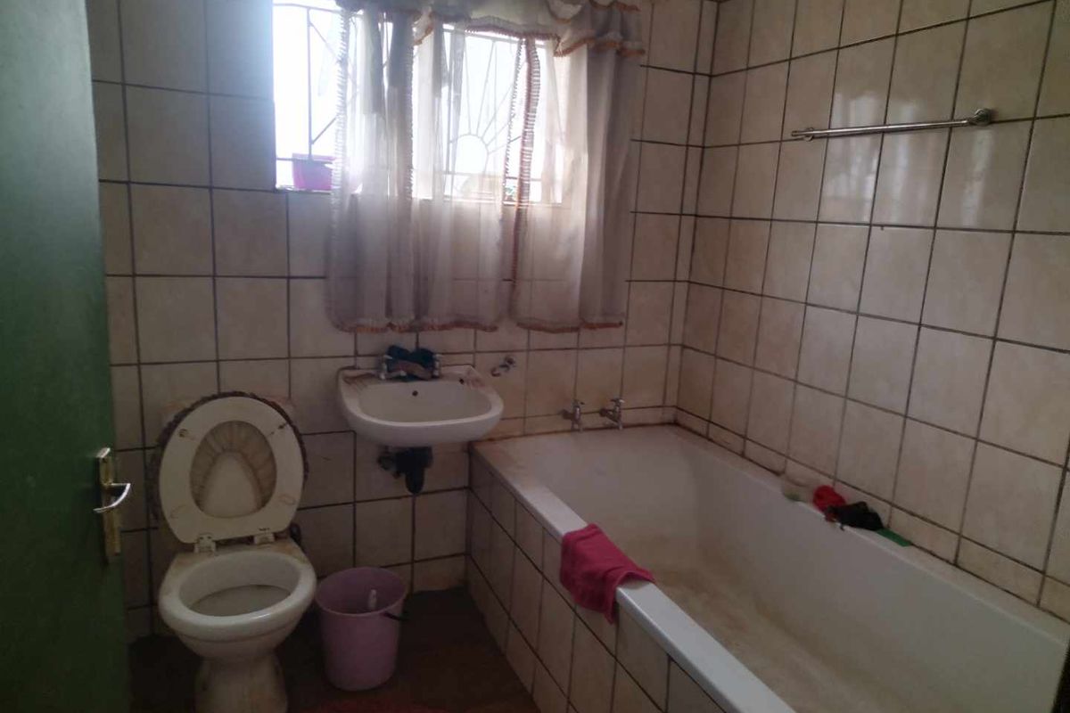 A dated bathroom featuring a standard bathtub, wall-mounted basin, and toilet with full-height wall tiling and security bars on the window.