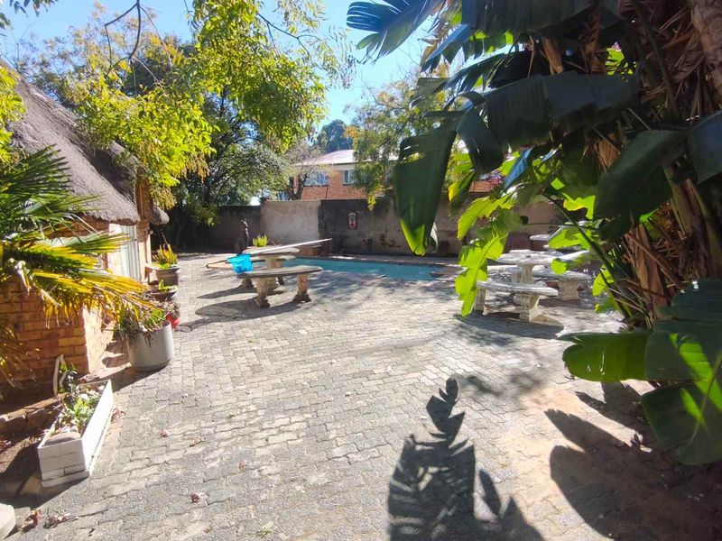 Outdoor pool area featuring a paved patio, swimming pool, and a thatched roof structure, surrounded by established garden greenery and boundary walls.