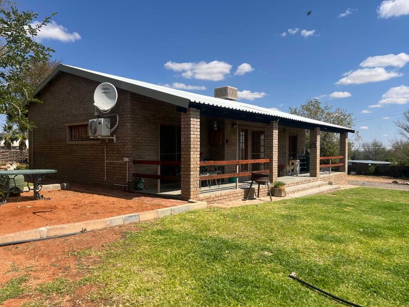 Rear view of a single-story brick house featuring a covered patio, corrugated metal roofing, and established garden space.