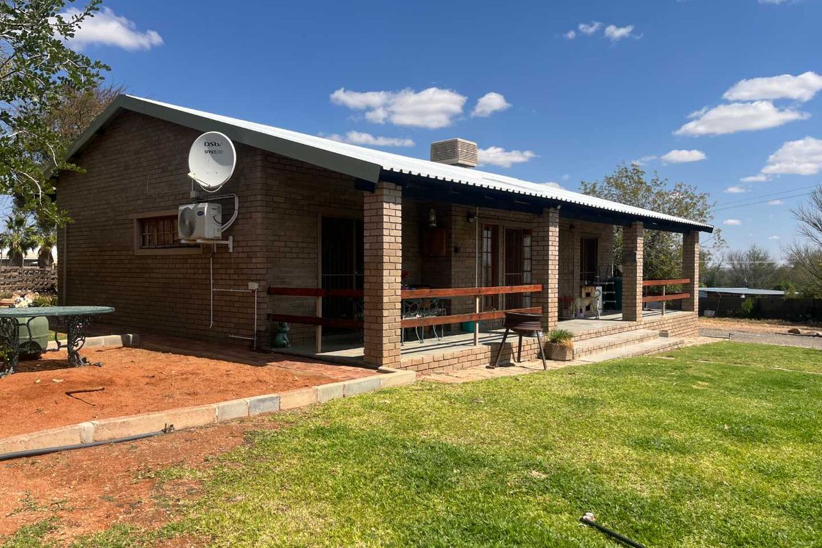 Rear view of a single-story brick house featuring a covered patio, corrugated metal roofing, and established garden space.