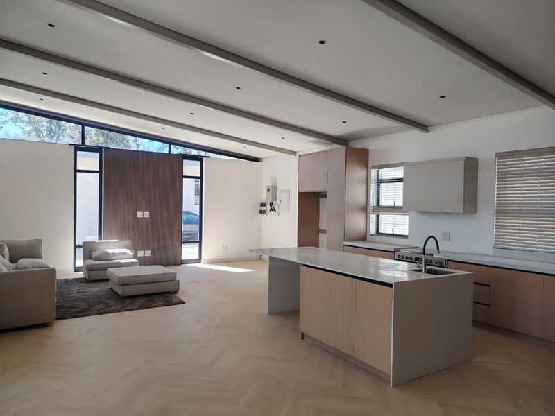 Modern open-plan living and kitchen area featuring herringbone flooring, exposed ceiling beams, a central island with sink, and large windows providing ample natural light.