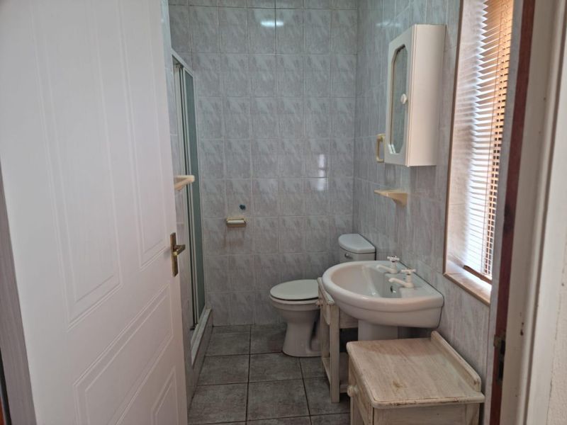 A functional bathroom featuring a shower enclosure, pedestal basin, and toilet with tiled finishes and natural light from a window with blinds.