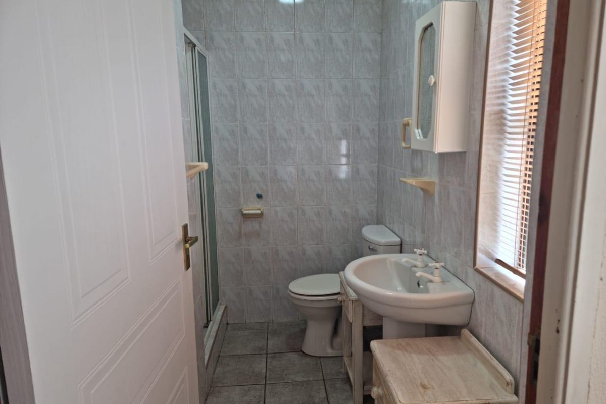 A functional bathroom featuring a shower enclosure, pedestal basin, and toilet with tiled finishes and natural light from a window with blinds.