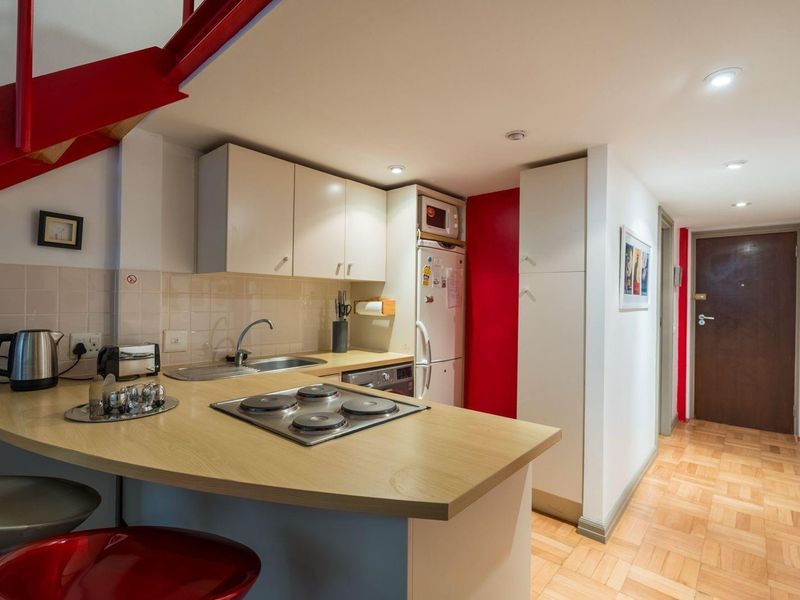 Compact kitchen featuring light wood-look countertops, built-in cabinetry, an electric hob, and tiled backsplash, situated adjacent to a hallway and staircase.