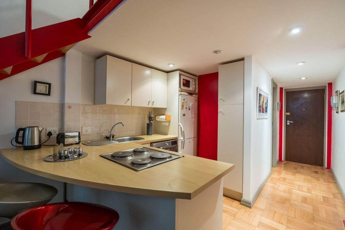 Compact kitchen featuring light wood-look countertops, built-in cabinetry, an electric hob, and tiled backsplash, situated adjacent to a hallway and staircase.
