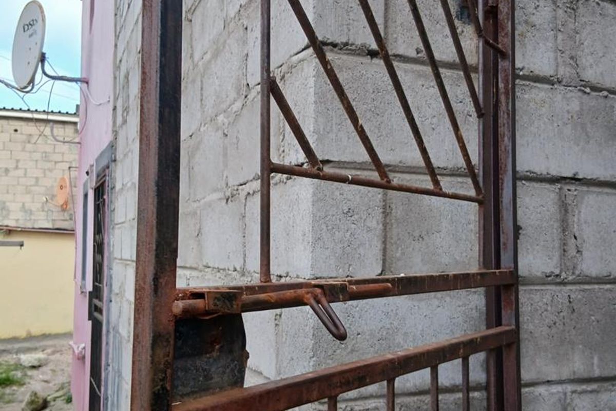 A view of a side or rear exterior area featuring a weathered metal security gate and unfinished concrete block walling.