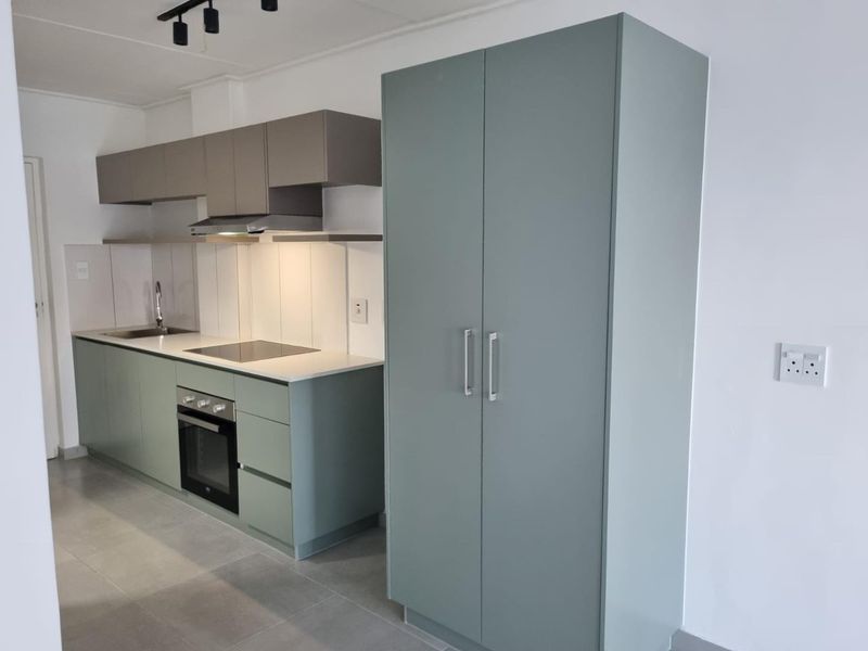 Modern kitchen featuring sage green cabinetry, integrated oven and hob, stainless steel sink, and neutral tiled flooring in a clean, minimalist space.