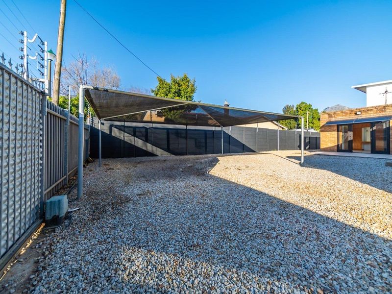 Rear exterior area featuring a gravel surface, shade sail structure, perimeter fencing with electric security, and access to the main building via sliding doors.