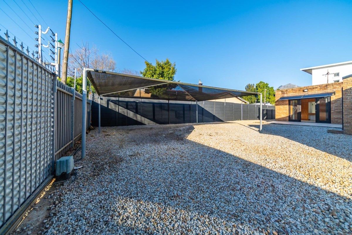 Rear exterior area featuring a gravel surface, shade sail structure, perimeter fencing with electric security, and access to the main building via sliding doors.