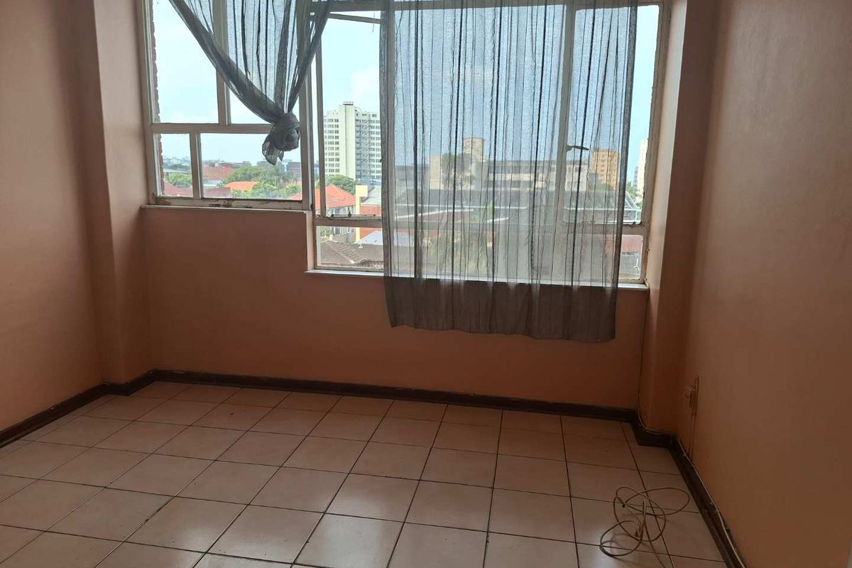 An empty bedroom featuring large windows with city views, tiled flooring, and peach-toned walls in need of cosmetic refurbishment.