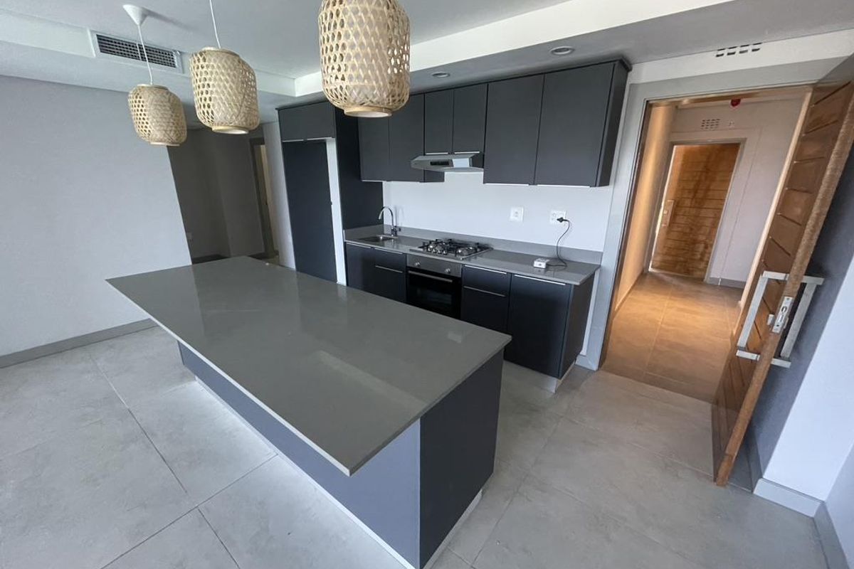 Modern open-plan kitchen featuring dark cabinetry, stone countertops, a central island, and integrated stainless steel appliances, set against neutral tiled flooring.