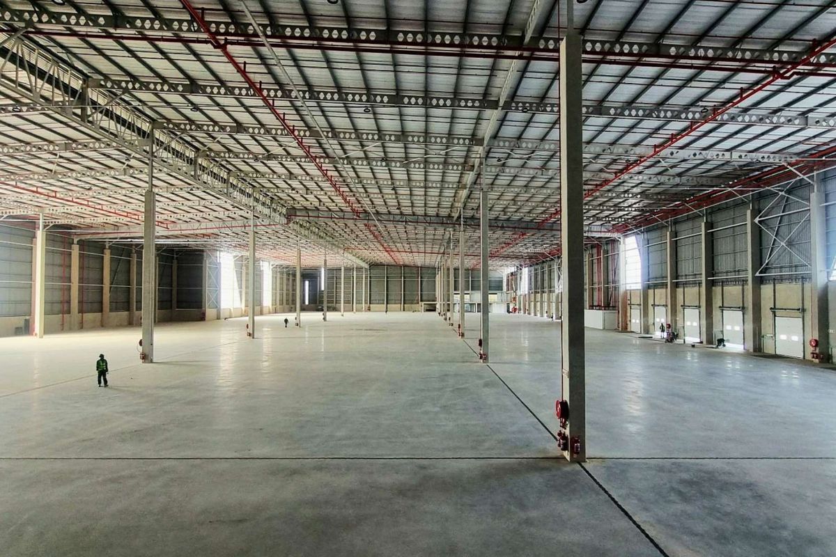 A vast, open-plan industrial warehouse space featuring high ceilings with exposed steel trusses, fire sprinkler systems, and polished concrete flooring.