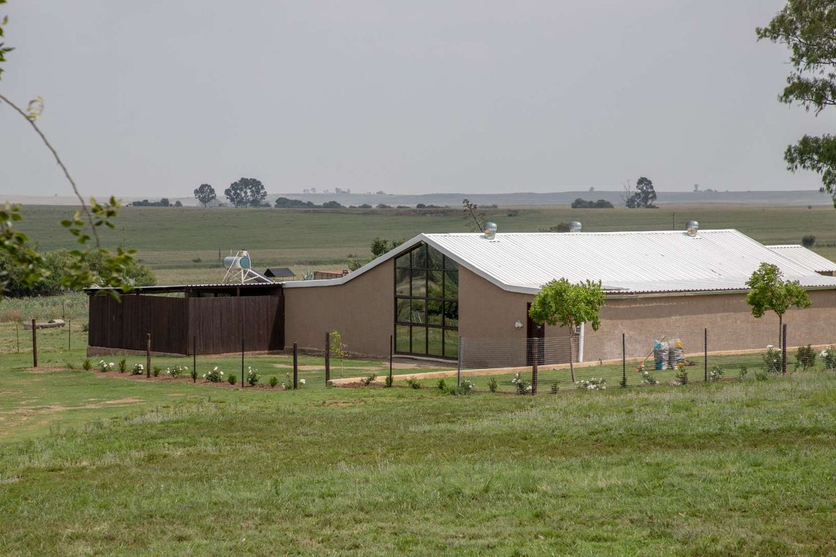 A rear view of a modern farm building featuring a large glass gable window, corrugated metal roof, and a fenced garden area.